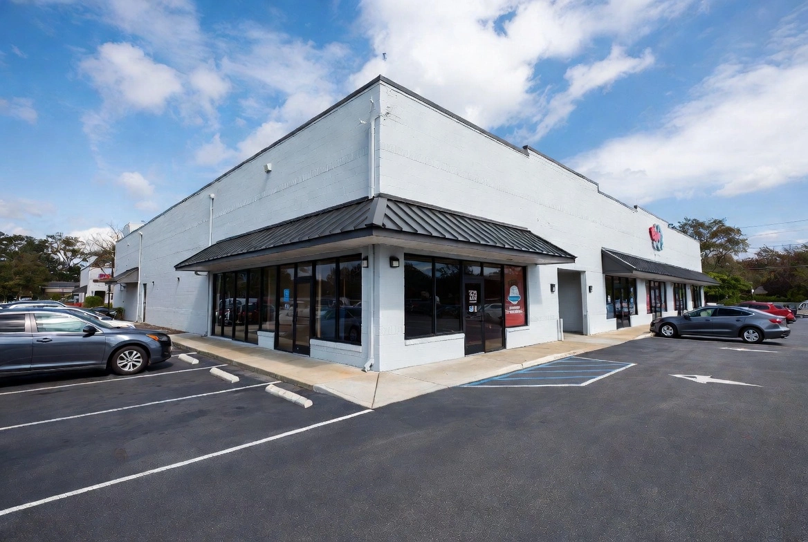 Corner view of a single-story white commercial building with black metal roof trim, large glass storefront windows, and parking spaces in front under a blue sky.