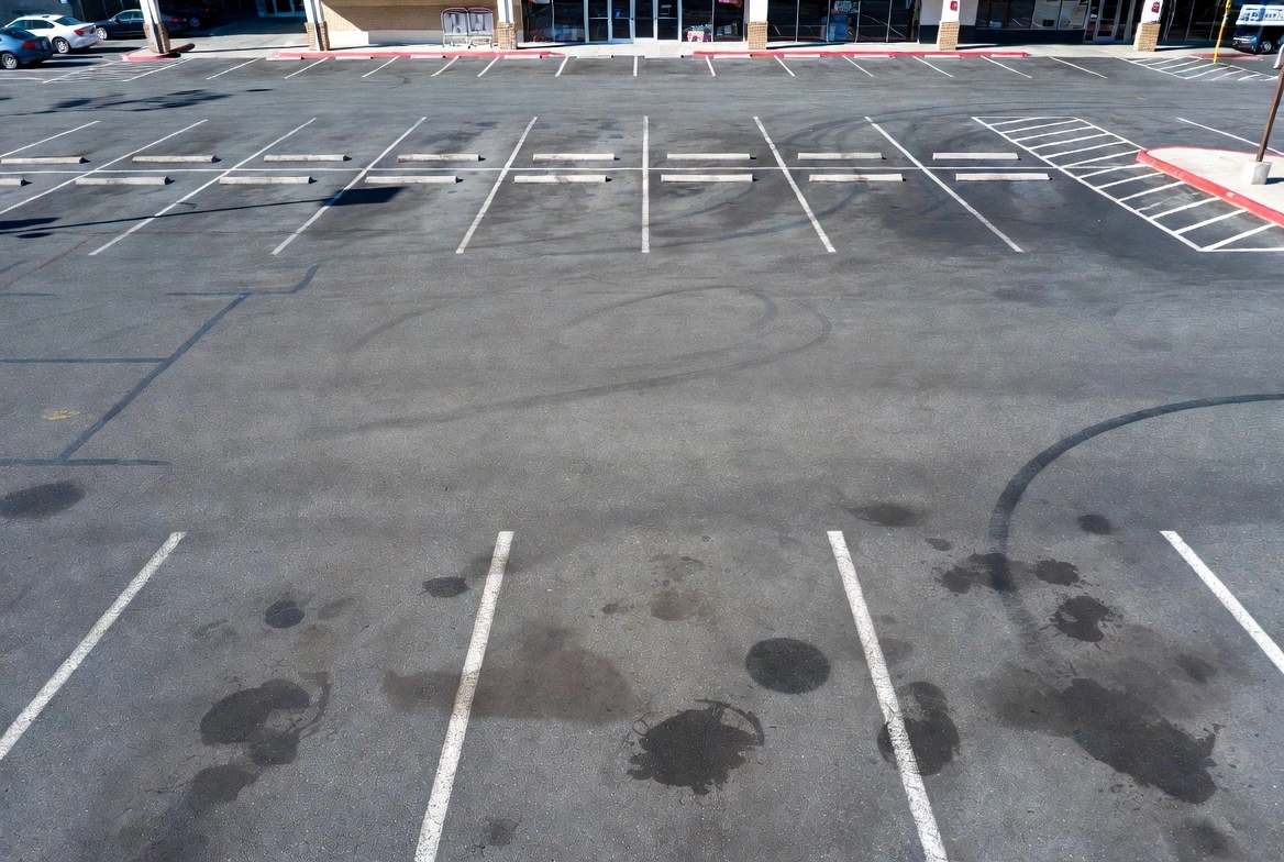 Close-up of a parking lot asphalt surface showing tire skid marks, oil stains, and dark spots on the pavement between parking lines.