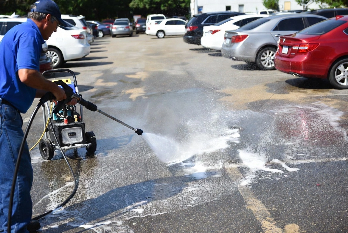 Man in blue shirt and cap operating a pressure washer on a parking lot, creating foamy water spray on the asphalt surface near parked cars.