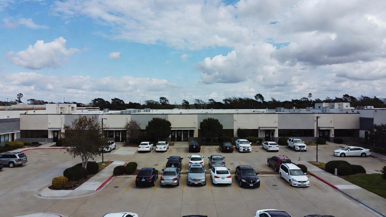 Aerial view of a commercial strip mall parking lot with many parked cars under a partly cloudy blue sky, beige and gray buildings with trees in the background.