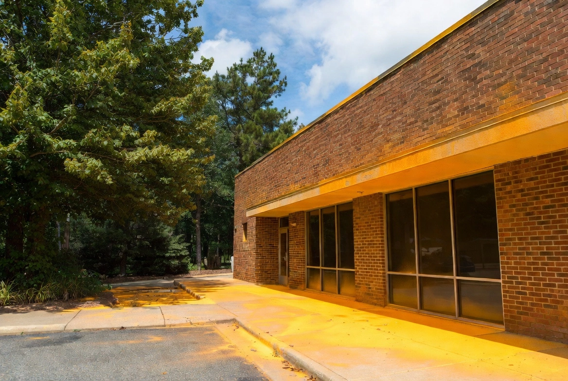 Brick single-story office building exterior with large windows, surrounded by tall pine trees under a partly cloudy sky